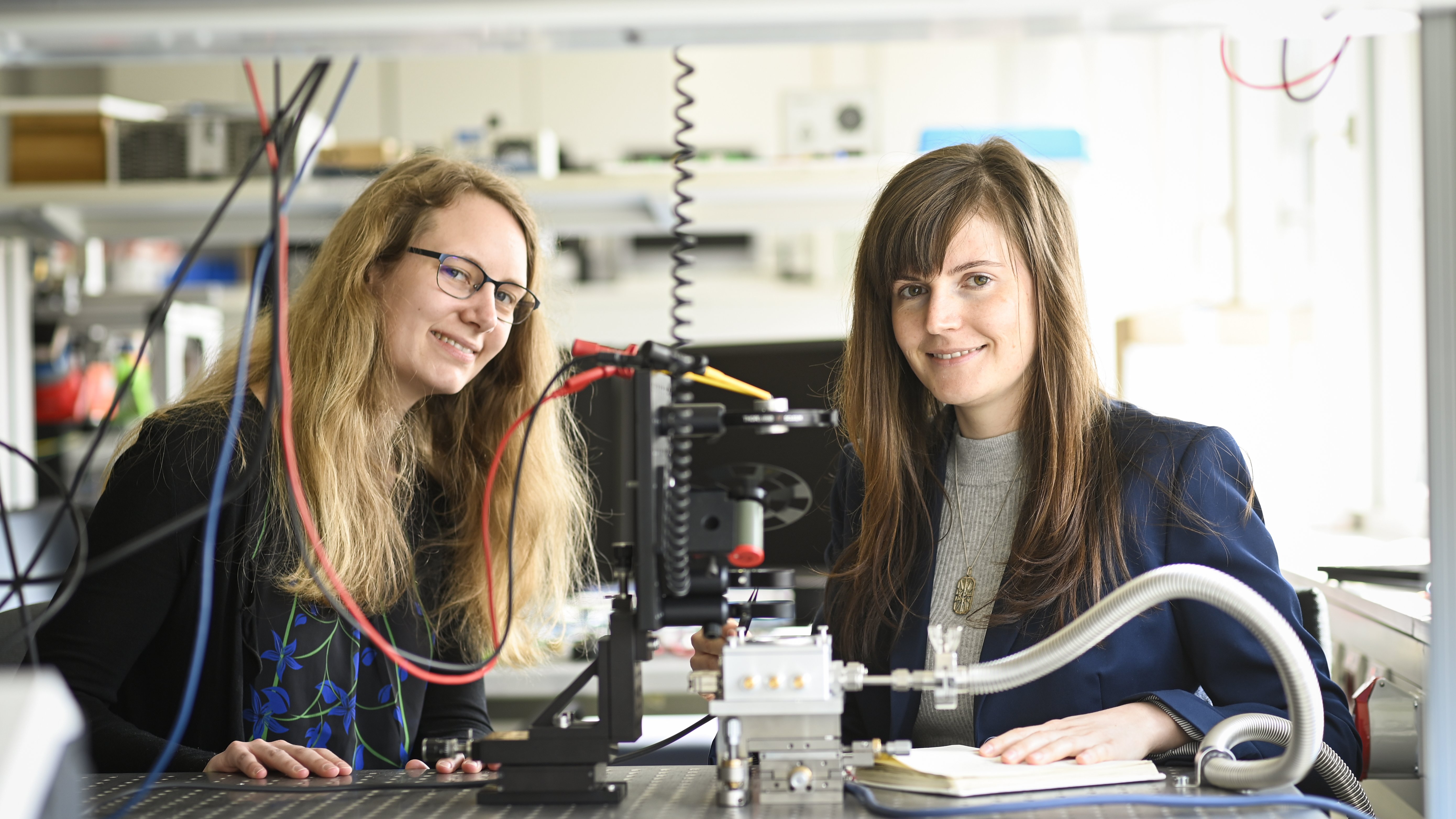 zwei Forscherinnen sitzen in einem Labor und lachen in die Kamera