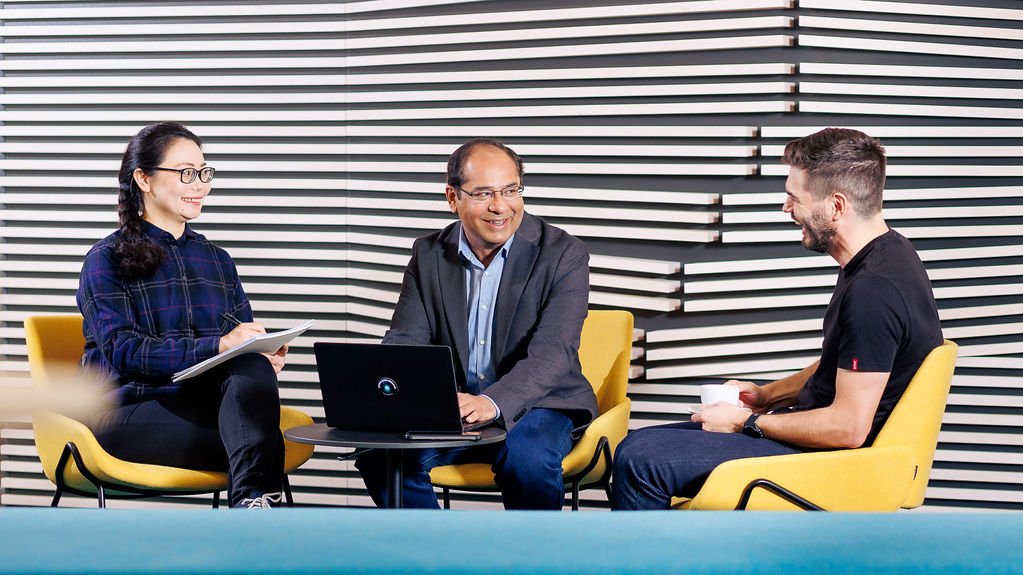 three people sitting on yellow office chairs, discussing something and laughing