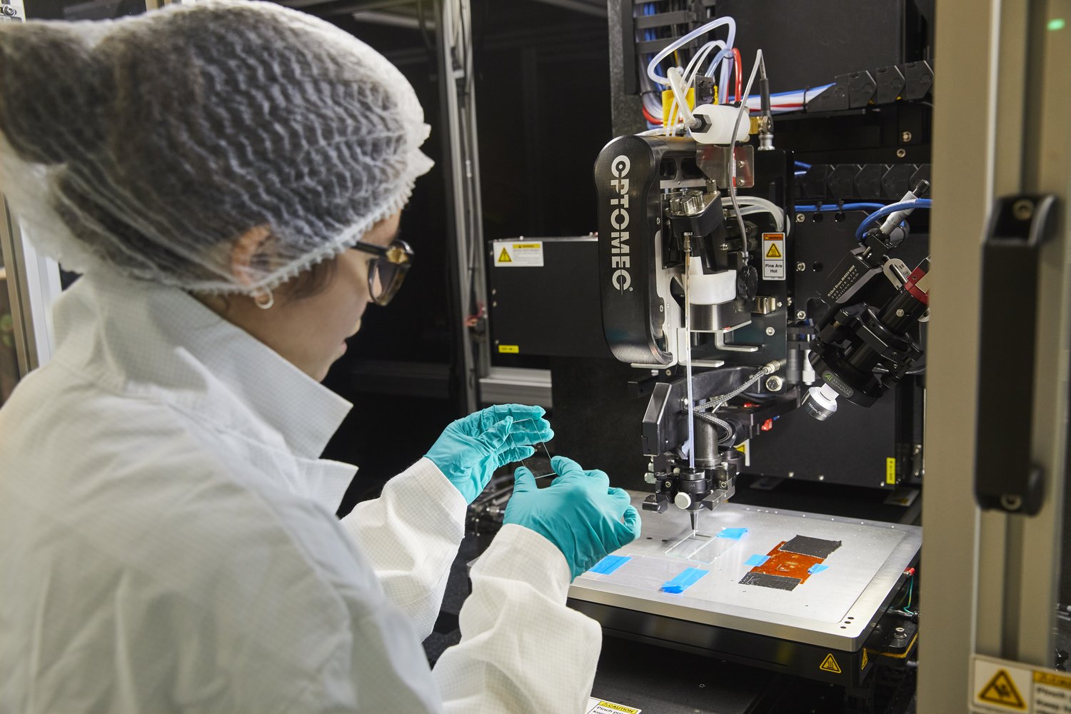 Emily Bezerra in protective clothing in the lab, looking at the Aerosol Jet Printer