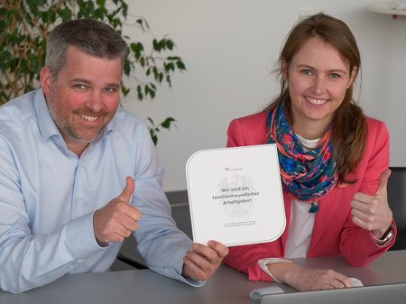 Executive Director Gerald Murauer and Emily Knes, Head of Human Resources, are excited about the re-certification as a family-friendly employer. They are sitting next to each other holding a certificate and giving a thumbs up