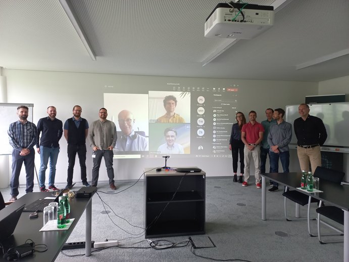 Gruppenbild beim Projektauftakt, 9 Personen stehen vor Leinwand, auf der drei weitere Personen online am Meeting teilnehmen