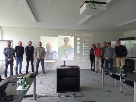 Gruppenbild beim Projektauftakt, 9 Personen stehen vor Leinwand, auf der drei weitere Personen online am Meeting teilnehmen