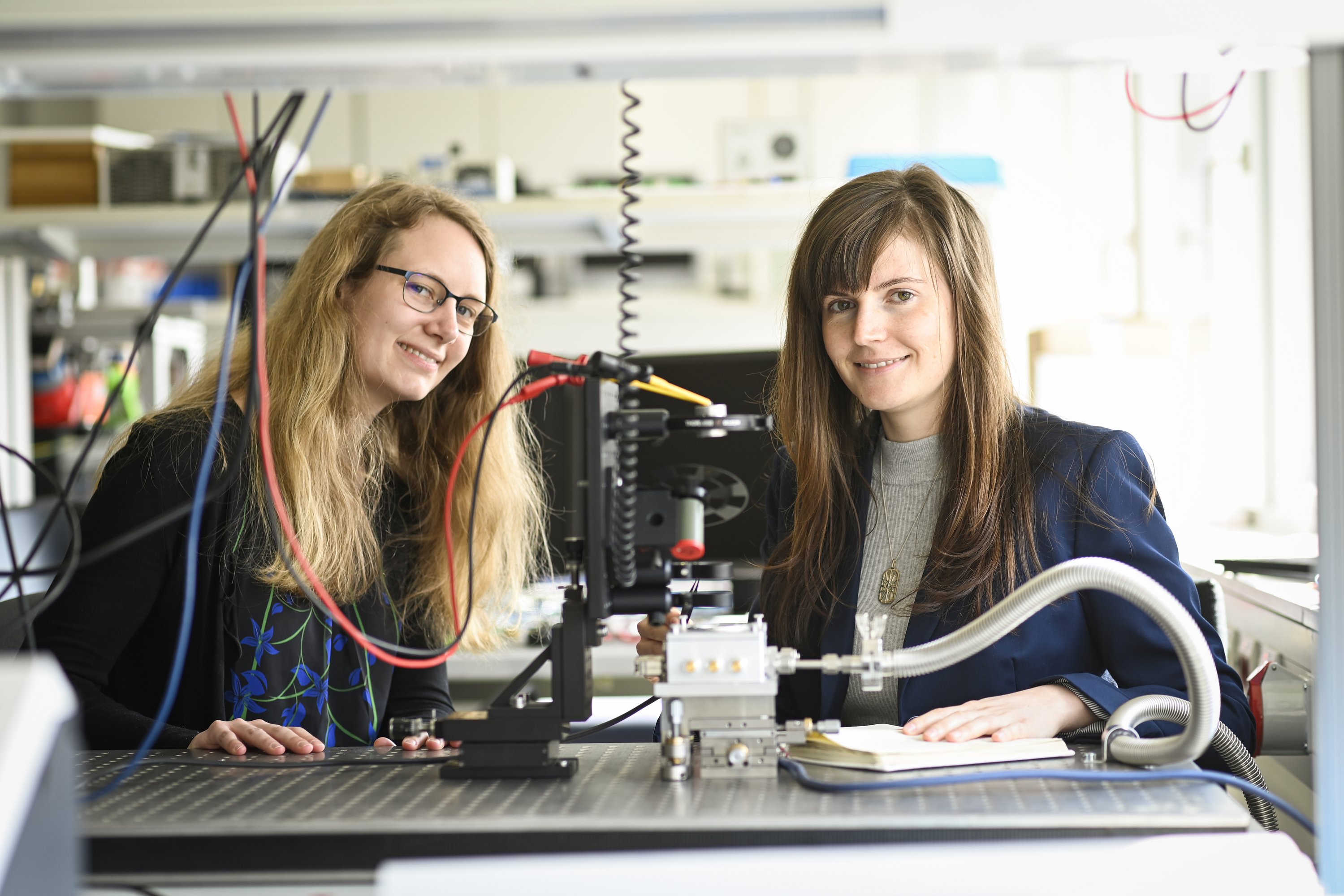 zwei Forscherinnen sitzen in einem Labor und lachen in die Kamera
